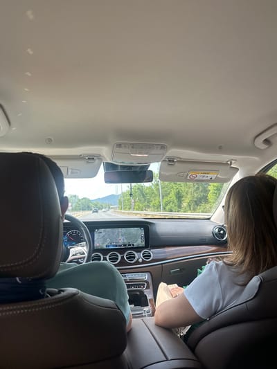 The image depicts the interior of a modern car from the perspective of a rear passenger seat, looking towards the front. The setting is daytime, with bright natural light suggesting clear, sunny weather.
Two people are present in the car. In the front passenger seat, a person with light brown hair, wearing a white t-shirt, is partially visible, seemingly holding a small, light-colored bag or wallet in their lap. The driver's head and shoulder, wearing a light blue or green shirt, are also visible, situated behind the steering wheel.
The car's interior features a sophisticated dashboard with multiple circular air vents and a large central display screen showing a navigation map. The time "12:11" is visible on the screen, and the dashboard also shows "km/h" and possibly "EQ," hinting at a Mercedes-Benz vehicle. A dashcam is attached to the rearview mirror. The passenger-side sun visor is lowered, displaying a yellow "A AIRBAG" warning label with universal safety pictograms, including warnings against placing a rear-facing child seat in the front.
Through the windshield, a scenic view unfolds, with lush green trees lining the road and prominent mountains in the distance under a bright sky. This landscape is consistent with the provided location context of Δημοτική Ενότητα Προμαχώνος, Greece, suggesting a journey through a picturesque, possibly mountainous region. The overall scene conveys a sense of a road trip or leisurely drive.