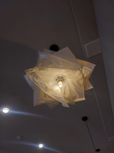 The image shows a close-up, low-angle shot of a unique, multi-layered white paper lampshade hanging from a dark ceiling. A bright incandescent lightbulb is visible in the center of the lampshade, illuminating the translucent material from within. Several smaller, circular lights are embedded in the ceiling in the background, along with a rectangular vent. The ceiling is a dark gray, textured material, and the overall lighting is dim, with the lampshade being the primary light source. There are no people or animals present in the image. The location is not specified in the image, but it is described as being in the city of Kyzylorda, Kazakhstan.