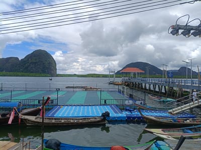 The image depicts a floating village scene with a distinctive football field on a pontoon. Several traditional longtail boats are moored in the foreground, their wooden hulls weathered by the sea. Some boats are decorated with colorful fabric. In the background, a long pier extends over the water, leading to a covered pavilion with a red roof.

The scene is set against a backdrop of lush green, rocky islands that rise dramatically from the calm, dark water. The sky is partly cloudy, with a mix of bright patches and heavier gray clouds, suggesting a day with variable weather. The time of day appears to be afternoon, given the lighting conditions.

A few people are visible on the pier, one man in a white shirt and shorts walking up a ramp. The overall atmosphere is serene, with a sense of local life and community evident in the floating structures and the boats. The presence of the football field on the water highlights the ingenuity and unique lifestyle of the inhabitants. Some indistinct text is visible on a blue sign on the pier and on a white banner on the floating football field. The location is Ko Panyi, Thailand, known for its stilted houses and floating facilities.