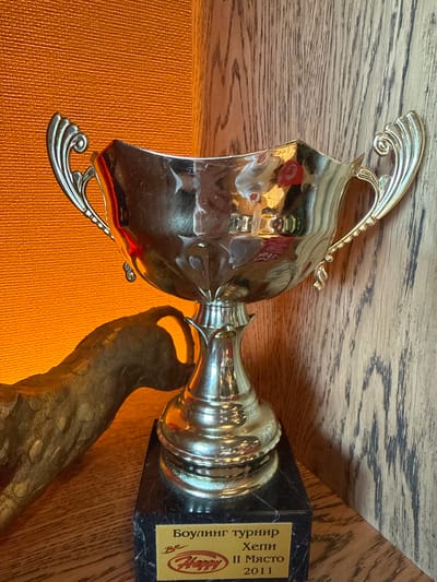 The image displays a gold-colored trophy, captured from an overhead perspective and oriented sideways. The trophy features a shiny, reflective cup with intricate, scroll-like handles on both sides. It is mounted on a dark, possibly marble, rectangular base.
A golden plaque is affixed to the base, containing text in Cyrillic script. The visible text reads "Боулинг турнир" (Bowling tournament), "Хепи" (Happy), "II Място" (2nd Place), and the year "2011". A stylized logo, likely for "Happy" and incorporating a smiling face, is also visible at the bottom of the plaque.
The trophy rests on a light brown wooden surface, possibly a shelf or table, against an orange, textured background. A blurred, dark, irregularly shaped object or shadow is partially visible beneath the cup of the trophy. The scene is an indoor still life, commemorating an achievement from a bowling tournament in 2011 where the participant earned second place. Given the location context of Radinovo, Bulgaria, and the Cyrillic text, the trophy is from a Bulgarian event.