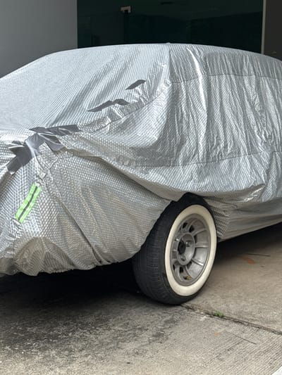 An aged car covered in a silver fabric car cover is parked on a concrete surface. A portion of the car's wheel, featuring a white-walled tire and a five-spoke silver rim, is visible on the right side of the frame. The car cover has several gray fabric tape strips attached to it, securing the cover. A small section of a bright green reflective strip is also present on the left side of the cover. The background shows a dark-colored wall and a sliver of a doorway or opening. The scene is set outdoors, in what appears to be a parking area or garage, possibly in Ban Tong Kai, Thailand, as indicated.
