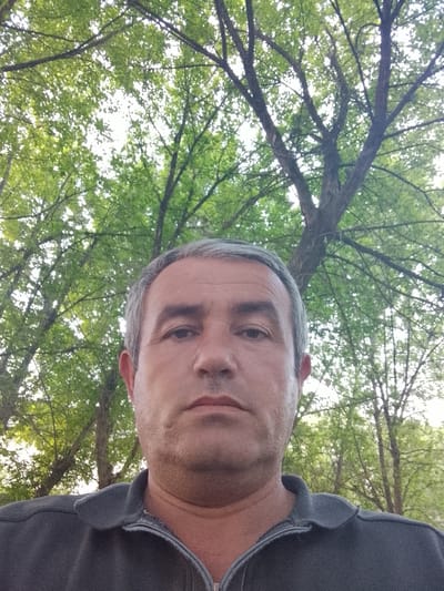 A middle-aged man is the primary subject of the image. He has gray hair and is wearing a dark gray zip-up polo shirt. The background is filled with trees featuring green foliage. The photograph was taken outdoors. The exact location in Syrdarya, Uzbekistan is not discernible from the image.