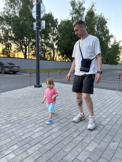 An adult male and a toddler are walking on a paved walkway. The male is wearing a white t-shirt, black shorts, white sneakers, and a black shoulder bag with "BOSS" written on it. He has tattoos on his left leg and is wearing a smartwatch. The toddler is wearing a pink t-shirt and blue and white plaid shorts with blue sneakers. They are on a brick-patterned walkway with bollards and a street sign in the background. Behind the walkway is a parking lot with a dark sedan and a grey fence. Trees are visible behind the fence. The sky is a gradient of yellow and blue, suggesting it is sunset or sunrise. The scene appears to be an outdoor public space, possibly a park or a parking lot area in the city of Kraskovo, Russia.
