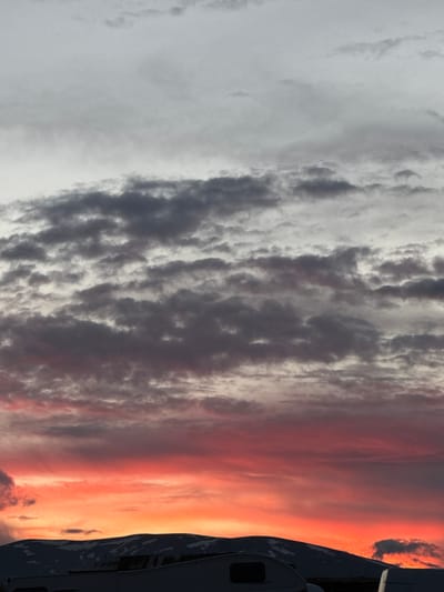 The image shows a sunset sky with clouds. The left side of the sky is grey, while the right side is colored with orange and red hues. A portion of a building or structure is visible on the right side of the image, with windows. There is no indication of people or actions. Anipemza, Armenia is not evident in the image.