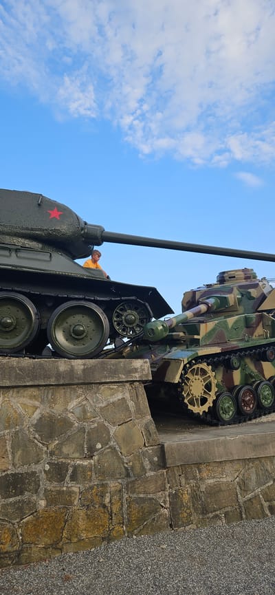In Svidník, Slovakia, two military tanks are displayed on a stone embankment. The tank on the left, painted dark green with a red star, has a young boy in an orange shirt sitting on its hull. The tank on the right is camouflaged in greens and browns. The sky above is blue with scattered white clouds. The embankment is constructed of large, irregularly shaped stones. The foreground consists of gravel.