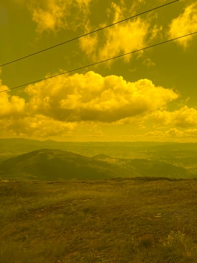 The image shows a mountainous landscape under a yellow-toned sky with dramatic clouds. Three parallel dark lines, likely power lines or cables, stretch across the upper portion of the frame. In the foreground, a grassy meadow slopes downwards, with a dirt path visible. Beyond the meadow, rolling hills covered in dark green vegetation recede into the distance, with layers of mountain ranges visible on the horizon. The overall impression is one of a remote, natural environment. The scene is devoid of any people or discernible structures, aside from what might be faint hints of buildings in the far distance. The coloration suggests an unusual atmospheric condition or post-processing effect. The location in Chorna Tysa, Ukraine, places this scene within the Carpathian Mountains.