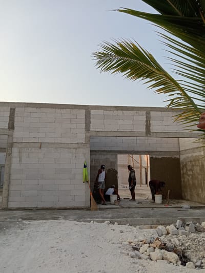 The image depicts a construction site with four individuals engaged in work. The structure appears to be a building under construction, with walls made of white aerated concrete blocks and concrete pillars. In the foreground, a pile of rubble and construction debris is visible on a sandy ground.
Three individuals are actively working. One person, wearing a white tank top and patterned shorts, is crouched down next to a white bucket, seemingly mixing materials or preparing them for application. Another individual, wearing a dark hooded sweatshirt and a cap, stands and appears to be overseeing or taking a break. A third person, wearing a brown shirt and dark pants, is bent over a white bucket with a shovel, also appearing to be working with materials. A fourth individual, wearing a white tank top and patterned shorts, is partially visible on the far left, standing near a broom and a bright green and yellow vest hanging on the wall.
The environment suggests an outdoor setting, with a clear sky above, tinged with the soft light of what could be dawn or dusk. Palm fronds are visible in the upper right corner of the image, indicating a tropical or subtropical location. The ground surface is a mix of sand and concrete. The building's unfinished state, with exposed blockwork and areas of plastered wall, points to ongoing construction activity.
While the prompt mentions "the city of Stiaji, Indonesia", there are no specific visual cues within the image that definitively identify this location. The presence of palm trees and the general construction style are consistent with many tropical regions, but without further information, a precise localization to Stiaji cannot be made solely from the visual content.