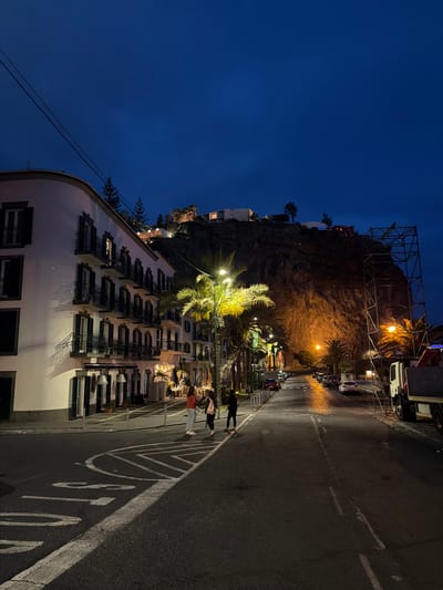 The image is a vertical, nighttime shot of a street scene in Ponta do Sol, Portugal. On the left, a multi-story white building with numerous black-shuttered windows is lit up, with a tall palm tree illuminated in front. People are seen along the street, some appearing to be dining outdoors. Behind the building and across the street, there's a rocky cliff illuminated with lights and what appears to be a scaffolding structure on top. A road with a painted stop sign runs along the right side, and several parked cars are visible further down the street. The sky is a dark, clear blue.