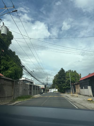 The image shows a street in San Rafael, Costa Rica, at the coordinates 9.936 latitude and -84.154 longitude. The street is paved and has two lanes. There are houses on both sides of the street. There are trees and bushes along the street. The sky is partly cloudy. The street is empty except for a white van in the distance. There are many power lines running along the street. There is a black gate on the left side of the image. There are several trees in the distance. The image is taken from a car, looking down the street. There are yellow lines in the middle of the street. There are utility poles on either side of the street. The image is taken from a vehicle looking down the street. There are some trees in the distance. The image is in focus.