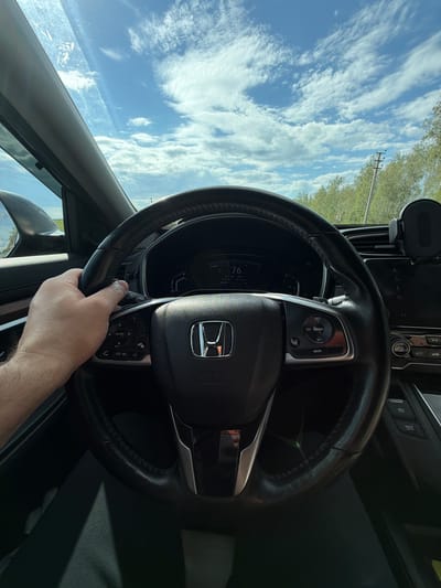 This media file shows a first-person perspective from inside a car, looking at the steering wheel and dashboard. A person's hand is visible gripping the steering wheel. The car's interior is dark, with a focus on the black leather steering wheel which has various control buttons on it. The dashboard also shows a digital display with the number "76" prominently displayed, likely indicating speed.
Through the windshield, a bright blue sky with white, wispy clouds is visible, suggesting it's daytime and the weather is clear. The background shows a hint of green foliage and a utility pole, indicating an outdoor setting, possibly a rural or suburban area. The angle of the sunlight suggests it might be mid-morning or afternoon.
There is visible text on the dashboard and steering wheel controls, including "RES", "LIM CANCEL", "SET", "MAIN", "ENTER", "P", "BRAKE", and "HOLD". The overall scene conveys the mundane act of driving. The time of day appears to be during daylight hours with clear skies.