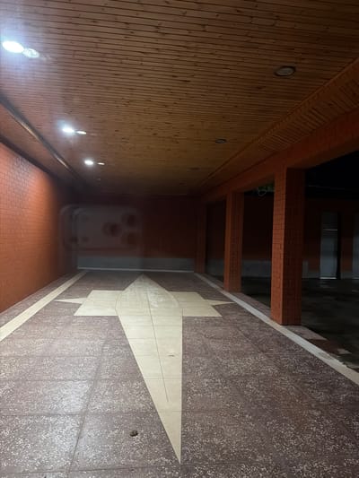 The image depicts an outdoor covered walkway or passage with brick walls and columns. The ceiling is made of wooden planks, and recessed lighting illuminates the space. The floor is tiled with a patterned terrazzo material, featuring a large arrow-shaped design in lighter tiles. On the left, a brick wall is visible, and on the right, there are brick columns supporting the roof structure. The walkway leads into a darker, seemingly open area or another building. There are no people or animals visible in the scene. The overall impression is of an architectural space, possibly part of a public building or a protected walkway in Kurchaloy, Russia.