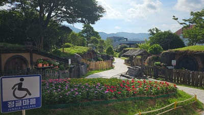 The image depicts a scenic view of the "Jungle Village" in Geoje-si, South Korea. In the foreground, a prominent blue and white sign features a wheelchair accessibility symbol and Korean text, indicating "Jungle Village." This sign is situated in front of a vibrant flower bed brimming with pink, purple, and red blossoms, bordered by lush green grass. A weathered wooden fence runs along the edge of the flower bed.
The path, paved with concrete, curves gently through the scene. To the left of the path, hobbit-like dwellings with rounded wooden doors are partially built into grassy mounds. Potted plants adorn the ledges of these structures. To the right, another hobbit-like dwelling with a rounded door and brickwork is visible, also covered with grass. A rustic wooden well with a stone base stands near the path, with a wooden shelter over it.
Wooden fences enclose various sections of the village, adding to the rustic charm. Beyond the village, trees, green hills, and distant mountains under a partly cloudy sky form the backdrop. A modern building with a glass facade can be seen in the distance. The overall atmosphere is serene and picturesque, suggesting a recreational or tourist area designed to evoke a whimsical, nature-integrated theme.