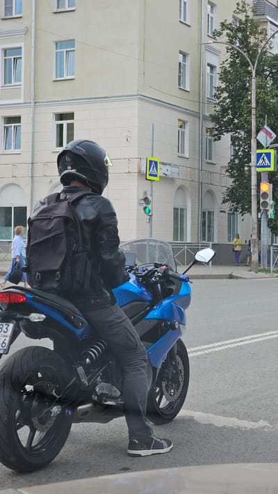 A person wearing a black helmet and a black leather jacket with a black backpack is sitting on a blue motorcycle at an intersection in the city of Kamensk-Uralsky, Russia. The motorcycle is facing away from the camera, with its rear tire visible in the foreground. The person is wearing dark pants and dark sneakers. In the background, there is a light-colored building with several windows. Across the street, there are traffic lights and pedestrian crossing signs. A woman in a yellow shirt and purple pants is standing on the sidewalk. The scene appears to be a city street on a cloudy day.