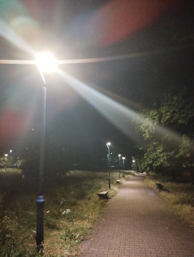 The image depicts a paved pathway in a park at night, illuminated by streetlights. A prominent streetlight is visible in the foreground, casting a strong beam of light across the scene. Several other streetlights are scattered along the pathway, with a few park benches placed on either side. Trees with dense foliage are present, and the ground is covered with grass and some fallen leaves. The overall setting appears to be a public park area within the city of Ruda Śląska, Poland, likely in the vicinity of a residential or recreational zone given the presence of park amenities. No people are visible in the image.