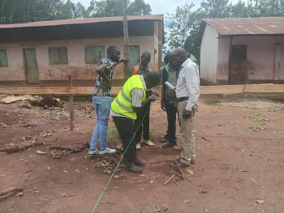 Five men stand on a dirt lot in Idakho South ward, Kenya, near two buildings with rusted metal roofs. One man in a yellow safety vest and black boots leans over, examining a green string stretched along the ground. Another man in a checkered shirt and jeans rests his arm on a wooden beam. Two other men observe, while the fifth man, wearing a white striped shirt and khaki pants, holds papers and a cell phone. The ground is covered with dirt, leaves, and small branches.