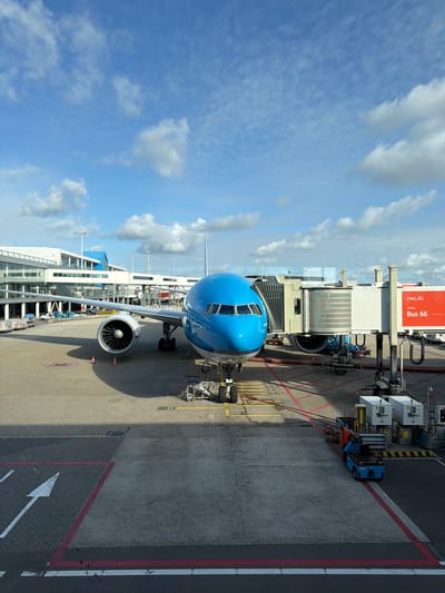 An aerial view shows a blue and white KLM airplane parked at a gate at Schiphol Airport in the Netherlands. The plane is connected to a jet bridge and is surrounded by airport vehicles and equipment. The sky is partly cloudy, with the sun casting shadows on the tarmac. The airport terminal building is visible in the background.