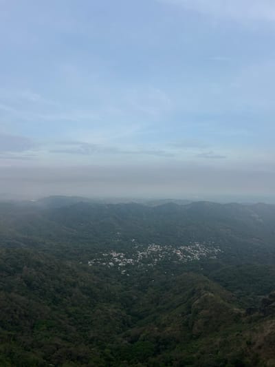 An aerial shot of the municipality of Planes de Renderos, El Salvador, shows a hillside covered in vegetation and scattered buildings. The sky is hazy, with clouds obscuring visibility.