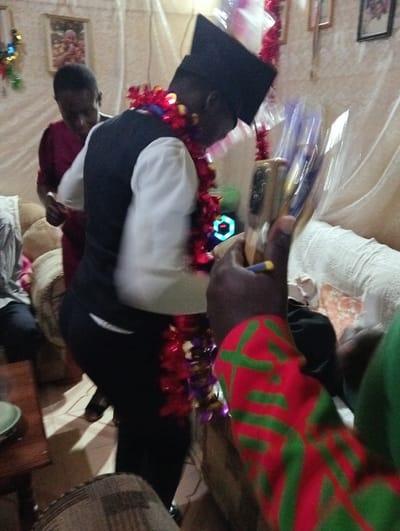A person wearing a graduation cap and a festive garland bows slightly, possibly receiving an award. Another person stands behind them, their attention directed downwards. In the foreground, a hand holds several objects, potentially pens or markers. The setting seems informal, possibly indoors with furniture and decorations visible in the background. One individual wears a red and green patterned sleeve.