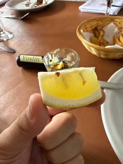 A hand holds a slice of lemon against a restaurant table. The lemon slice is bright yellow with a thick white rind. The table has various items including a woven basket filled with what appears to be fried potatoes, a small glass, and cutlery. "Yoslin" is printed on a small black bottle. The lighting is bright and even, illuminating the scene.