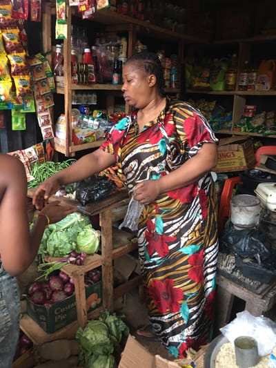 The image captures a vibrant market scene in Isieke, Nigeria, showing a woman attending to a customer in a small, well-stocked stall. The photograph is oriented sideways.
The main subject is a woman with braided hair, wearing a striking, colorful dress adorned with large red flowers and a black-and-white zebra-like pattern. She is leaning forward, reaching down towards the hands of another person, whose lower torso and arms are partially visible in the bottom right corner, suggesting an interaction or transaction.
The setting is a busy, perhaps cluttered, market stall or small shop. Wooden shelves line the background, filled with various packaged goods, including sachets, bottles, and cans. On the ground level, fresh produce such as large green cabbages, red onions in wooden crates, and green beans are displayed. A large metal bowl containing grains, with a metal measuring cup resting inside, is visible in the upper right. Other items include a box of "KING'S VEGETABLE OIL" and various household objects. The lighting suggests it is daytime, possibly within a covered or shaded area.
Visible text on products includes "LAZIZ," "INGS," "Custard Pie," "ARSOA," "WAO," and "KING'S VEGETABLE OIL FOR COOKING AND SALADS." The woman appears engaged and focused on the exchange with her customer.