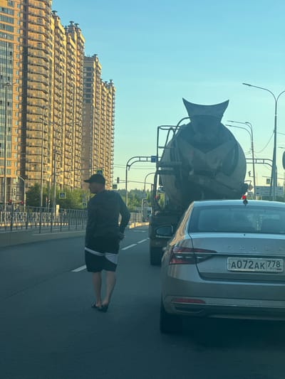 In Pargolovo, Russia, on a sunny day, a man wearing a dark hooded jacket, dark shorts with white trim, and dark sandals walks on a road. He is facing away from the camera, looking towards a large concrete mixer truck. The truck is in motion, as is a silver Skoda sedan in the foreground, whose rear license plate reads "A072AK 778 RUS". In the background, tall, yellow residential buildings line the street, with balconies visible on each floor. Traffic lights and streetlights are present, along with road signs. The sky is clear and blue. The scene appears to be a busy urban or suburban environment.