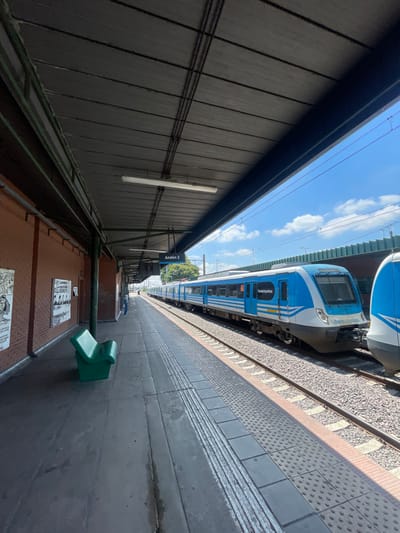 A blue and white train occupies the right side of the frame, parked at a station platform. The platform is grey with tactile paving. On the left, a dark grey wall runs parallel to the train. A brown, brick-like structure with posters attached sits beneath the wall, with a green object mounted atop. The sky, filled with white clouds, is visible between the train and the wall. An "Andén 2" sign is visible on the left side.