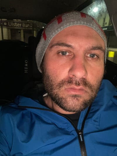 A man is in a car near an Ulster Bank in Newry, United Kingdom. He is wearing a gray beanie with red stripes, a black shirt with the word "HERO" on it, and a blue jacket labeled "Superdry". The time is likely nighttime.