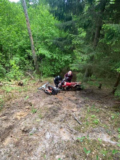 The scene depicts a wooded area in Парэцкі сельскі Савет, Belarus. Two dirt bikes and riders are situated on a steep, muddy incline within the forest. One motorcycle is red, while the other appears to be white. Both riders are wearing protective gear. The surrounding environment is characterized by lush green foliage, tall trees, and the uneven terrain of the forest floor. The conditions appear to be challenging for the dirt bikes, possibly due to the steepness of the slope and muddy ground.