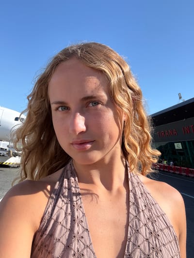 A young woman with blonde, wavy hair takes a selfie outdoors. She is wearing a light brown, patterned top. The background shows a bright blue sky and a building with the text "TIRANA INT" visible, suggesting an airport or similar transportation hub. The woman is looking towards the camera with a neutral expression. Sunlight is casting shadows on her face and clothing, indicating it is daytime. There's a part of an airplane visible at the bottom of the image. The overall impression is a casual, sunny day outing.