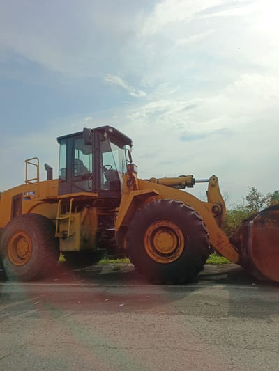 An all-yellow front-end loader, likely a Caterpillar 988K, is parked on an asphalt surface. The loader is positioned facing right, with its bucket lowered and resting on the ground. The sky above is partly cloudy, with a bright sun casting a glare towards the top right of the frame. The surrounding environment includes green foliage and trees to the right of the loader. The loader has large, black, treaded tires, a prominent cab with tinted windows, and yellow hydraulic cylinders extending from the body to the lifting arm. There are no people visible in the image. The location is identified as Nizhnii Atlyan, Russia.