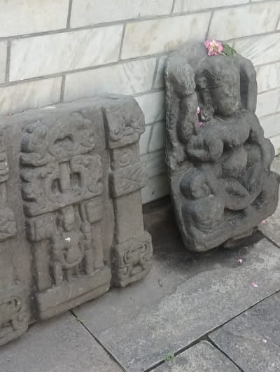 Two stone sculptures are displayed outdoors against a wall constructed of marble tiles. The sculpture on the left is a rectangular block with intricate carvings, including what appear to be faces and decorative elements. The sculpture on the right is a roughly shaped stone depicting a seated figure, possibly a deity or person, with delicate pink flowers placed on its head and fallen petals scattered around it. The ground is paved with large stone tiles, and a few small green sprouts are visible between the tiles. The scene is likely in a historical or religious site in Gojra, India.
