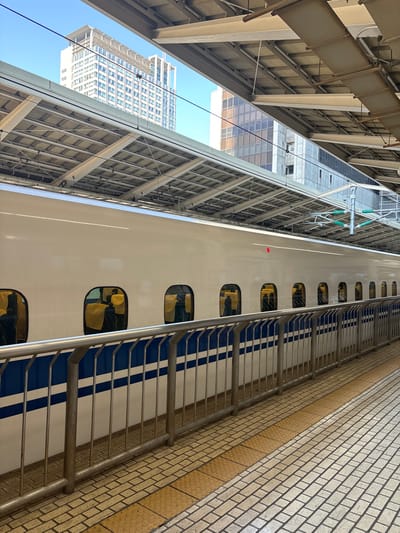 The image captures a white bullet train, part of the Shinkansen high-speed rail network, from a low-angle perspective. The train is parked beneath a steel structure that supports a nearby bridge or elevated walkway. The structure's metal beams and latticework cast a shadow on the side of the train. The train's blue and white striped exterior design is visible, with several windows revealing glimpses of the interior.
The train is situated at the coordinates 35.681, 139.768, which corresponds to a location in the Chiyoda ward of Tokyo. The surrounding area is a mixture of urban development and infrastructure. A tall, modern building with a glass facade appears in the background, suggesting a densely populated city environment. The image is taken from a vantage point that allows the viewer to see the train from a unique and slightly elevated position.