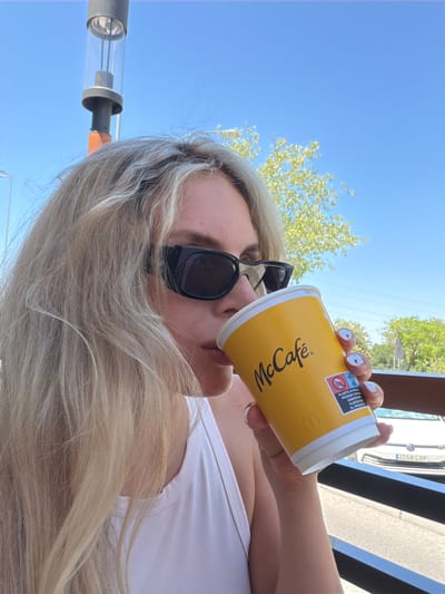 A woman with blonde hair and black sunglasses is drinking from a McCafé cup outdoors on a sunny day. She is wearing a white tank top. The background shows a clear blue sky, some trees, and a white car. There is also a lamppost visible in the upper left corner of the image. The setting appears to be a public outdoor area, possibly a cafe patio, within the city of Húmera-somosaguas-prado Del Rey, Spain, as indicated by the visible car license plate.