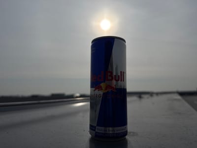 A Red Bull energy drink can is centrally positioned and upright on a light-colored, flat surface in Naaldwijk, Netherlands. The can is primarily blue and silver, featuring the red "Red Bull ENERGY DRINK" logo and two red bull motifs. Text visible at the base includes "Vitalizes body and mind" and "HIGH CAFFEINE CONTENT." The background consists of an overcast sky, with the sun appearing as a bright, distinct disc directly behind and slightly above the can, casting a strong backlight. A blurred horizontal line, possibly a railing or edge, extends into the distance on the surface. Indistinct, dark architectural or industrial forms are visible on the distant horizon. No individuals or discernible activity are present in the scene.