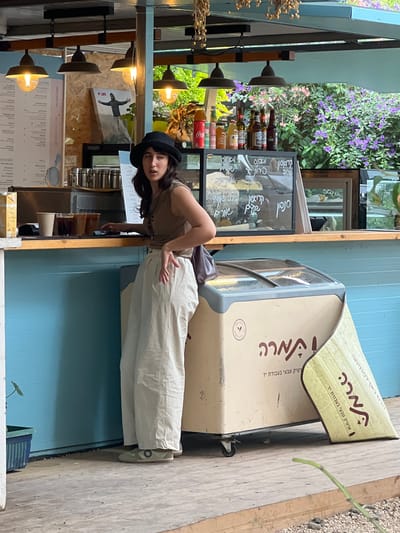 Here is a description of the image:
The image shows a woman in a hat standing in front of a building. The building has a menu on the wall, and there is a sign that reads "Mara, Artic Teva Beovdat Yad," which translates to "Mara, Natural Arctic in Crafting Hand," likely indicating a local artisan ice cream shop. The surrounding area appears to be a café or restaurant setting. The environment is partly obscured by various objects like shelves, drinks, and other decorative elements, and appears to be Jezreel Valley Regional Council, Israel.