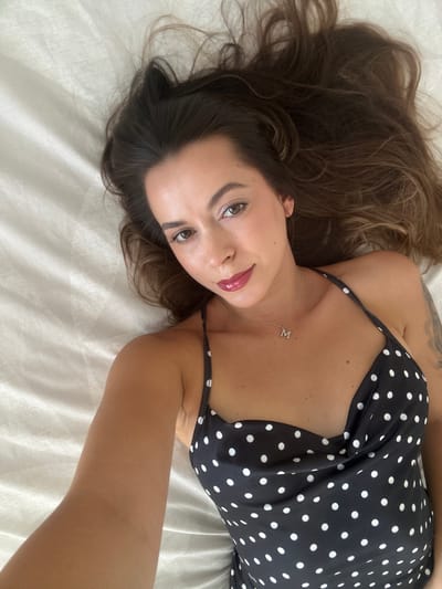A woman with long, dark brown hair is lying on a bed with white sheets. She is looking up at the camera, with her head tilted to the side. She is wearing a black and white polka dot dress and a silver necklace with a charm shaped like the letter "M". Her lips are painted with a dark pink lipstick. The lighting suggests an indoor setting, possibly a hotel room or bedroom in Torremolinos, Spain.
