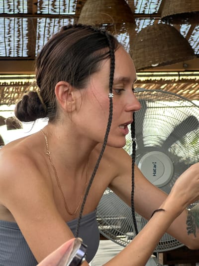 The image captures a close-up, profile view of a woman in a warm, bright setting, likely indoors or under a covered structure in Hà Giang 1 Ward, Vietnam. She is looking upwards and slightly to her left, with her mouth agape in an expression that suggests concentration or the need for breath. Her dark hair is partially styled into multiple thin braids, one of which has a small silver flower charm, and another a tiny silver ring. She wears a grey tank top and a delicate gold chain necklace, the clasp visible at the back of her neck.
Her arms are raised and crossed, with a prominent green and black tattoo visible on her upper left arm. Her right hand appears to be holding a clear plastic object, possibly a spray bottle or a small drinking bottle, while her left hand is near her head, seemingly engaged in adjusting her braids or hair.
A large grey Hatari brand standing fan is prominent in the background, its blades visible and in motion, indicating an effort to cool the area. The brand name "Hatari" is clearly visible, though reversed, on the fan's motor housing. The surrounding environment features intricate woven bamboo or straw lattice work, suggesting a rustic or traditional architectural style common in warm climates. The bright, natural light filtering through these structures indicates it is daytime.