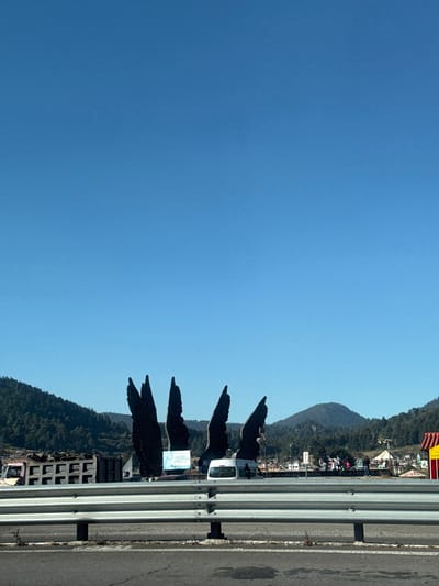 A sideways landscape view features a clear blue sky filling most of the frame. Along the right edge, a road with a guardrail runs parallel to the image's side. On the right, a mountainous terrain covered in trees and buildings is visible. Tall, dark silhouettes, seemingly decorative elements, are arranged along the roadside between the guardrail and the mountainous area. A white van is also parked along the roadside.