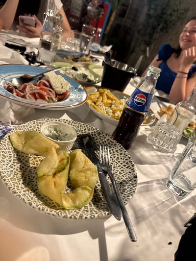 The image depicts a table setting at an outdoor restaurant in Kavala, Greece, likely during an evening meal. In the foreground, a plate with fried zucchini slices and a small dish of tzatziki sauce is visible. To the right, a Pepsi bottle and a glass suggest a beverage is being consumed. In the background, a woman in a blue top is smiling and looking towards the right. Other plates of food, including a salad with tomatoes and feta cheese, and a plate of french fries with cheese, are scattered across the table. Several glasses and bottles are also present. The overall scene suggests a relaxed and enjoyable dining experience.