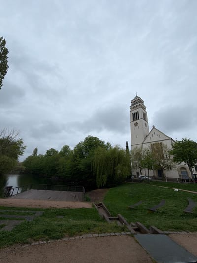 The image shows St. Johannes Nepomuk Church in Kehl, Germany, viewed from a waterside perspective. The church, a tall, white structure with a prominent clock tower, stands against a cloudy sky. Lush green trees and grass border the waterfront. A wooden platform or dock extends towards the water. The viewpoint is from a gravel pathway next to the waterfront. No people are visible.