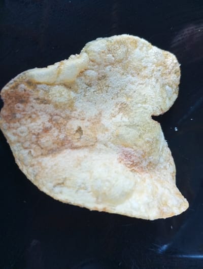 A single, irregular-shaped potato chip rests on a dark, reflective surface, possibly a plate or countertop. The chip is light brown and golden in color, with some darker, almost reddish-brown patches. Its surface appears slightly bumpy and textured, with a scattering of white powdery residue, suggestive of salt or seasoning. The background is uniformly dark and out of focus. There is no indication of any specific location within the city of Dymer, Ukraine, or any people, actions, or interactions.