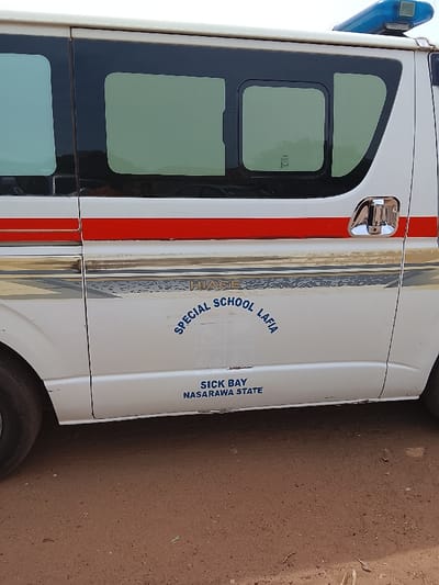The image shows a white van with a red stripe and gold lettering. The van has a "HIACE" emblem and "SPECIAL SCHOOL LAFIA" written on the side. The van also says "SICK BAY NASARAWA STATE" on the side. The van is parked on dirt ground. There are no people in the image. The coordinates provided place the image in a rural area of Nigeria, likely far outside of any city.
