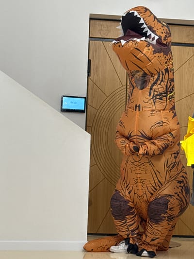The image features a person dressed in an inflatable T-Rex costume. The costume is orange with black markings resembling scales and stripes. The T-Rex has its mouth open, revealing white teeth. The person is wearing white sneakers with black laces. The costume is inflated, giving it a bulky and rounded appearance.

The scene appears to be indoors, with a wooden door or paneling visible in the background. The floor is a light-colored wood or laminate. To the left of the frame, a small rectangular device with a blue screen is mounted on a light gray wall. The screen displays text that appears to be "Google." A yellow piece of fabric is draped over the top of the T-Rex costume, partially obscuring the head.

The lighting suggests it could be daytime, but there are no clear external windows to confirm the weather. The overall impression is one of playfulness or a costume event. The orientation of the image is such that the T-Rex is leaning or standing in a slightly tilted manner.