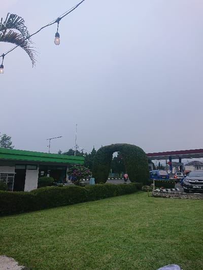 The image presents an outdoor scene located in Cibeureum, Indonesia, under an overcast sky. The foreground consists of a well-maintained green lawn, bordered by a dark green, trimmed hedge running horizontally across the mid-ground. To the left, behind the hedge, a single-story building with a white facade and a green roof is visible. Centrally, a large, U-shaped archway constructed from dense green foliage stands alongside a paved roadway. A person on a motorcycle, wearing a pink garment, is observed passing through this foliage archway. To the right, a gas station with a prominent red and blue canopy is active, displaying multiple fuel pumps. A blue van and a dark-colored SUV are present near the gas pumps. In the upper left quadrant, a string of two illuminated Edison-style light bulbs is strung across the frame, partially obscured by a palm frond. The background includes additional indistinct vehicles, utility poles, and a tall, slender mast.