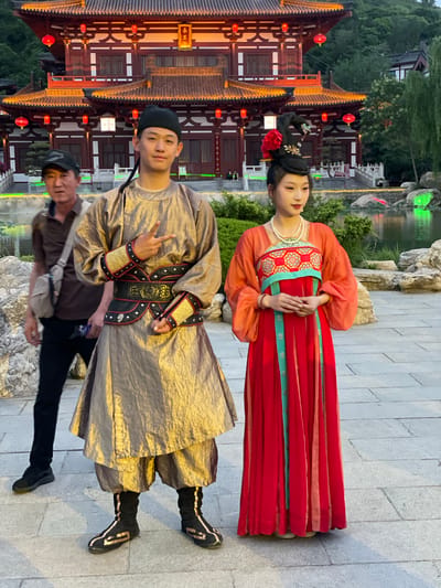 In Lintong District, China, a man and woman are dressed in traditional clothing and stand for a photograph near a pavilion with red lanterns. The woman wears a red and orange dress, and the man wears a gold garment. Another man stands nearby. A body of water and trees are visible in the background.
