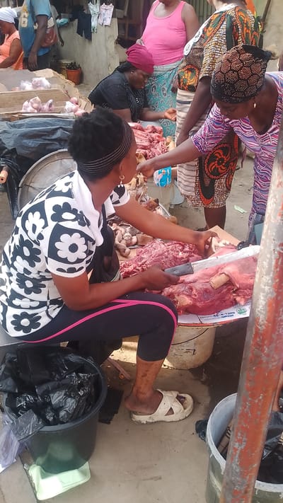 In a market in Warri, Nigeria, a woman in a white shirt with black floral patterns and pink-striped leggings sits behind a table covered with raw meat and cuts a portion with a large knife for a customer in a patterned shirt and headwrap. The butcher has a black headband and gold earrings. In front of her, to the left, is a black bucket filled with black plastic bags. At the next table over, another woman wearing a pink tank top and patterned skirt stands. A third woman with a red head covering is also seated, apparently selling chicken, near the pink-shirted woman. Another man in a blue shirt is standing near that woman, near a pile of fresh chicken. The market area appears busy with other vendors and customers. A metal support structure is visible in the foreground, to the right of the frame.