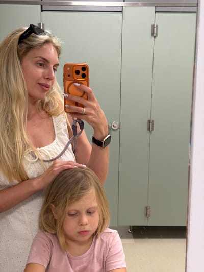 A woman and a young girl are reflected in a mirror, possibly in a restroom stall. The woman is on the left, holding a phone to take a selfie. She has blonde hair and is wearing a white, textured top and sunglasses. A ring is visible on her right hand, and she has a watch on her left wrist. The girl is on the right, leaning her head on the woman's arm. She has blonde hair and is wearing a pink t-shirt. The background consists of light green stall doors with metal hardware. The overall lighting appears to be artificial, suggesting an indoor setting during the day or evening. The reflection in the mirror shows the woman looking at her phone and the girl with her eyes closed, appearing relaxed or sleepy. There is no visible text in the image.