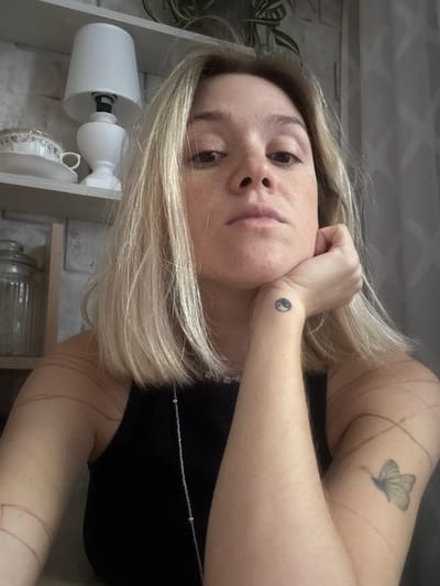 The media file is a slightly tilted, close-up selfie of a young woman with blonde hair. She is resting her chin on her hand, and her expression appears thoughtful or pensive. She has a small Yin and Yang tattoo on her left wrist and a butterfly tattoo on her upper left arm. She is wearing a black top with a delicate, beaded necklace. In the background, shelves are visible with objects including a white lamp, a decorative cup, and a glass jar. The setting appears to be indoors, possibly a home environment, with a hint of natural light. There is no visible text in the image. The overall mood is calm and introspective. The crops provide closer views of the woman's face, her tattoos, and some of the background objects.