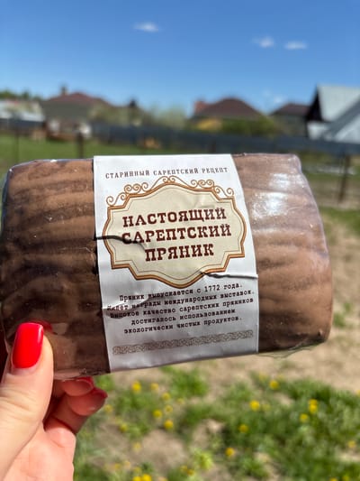 Here is a description of the image:
A hand with red nail polish holds a package of "Nastoyashchiy Sareptskiy Pryanik" (Genuine Sarepta Gingerbread). The label on the package indicates it is made according to an old Sarepta recipe and has won international awards. The text on the label is in Russian. The background appears to be outdoors, with grass and a blurry building visible under a blue sky. Based on the OCRed text, this image was likely taken in or relates to the city of Малые Кабаны, Russia, given the reference to Sarepta, the former German settlement near present-day Volgograd, Russia.