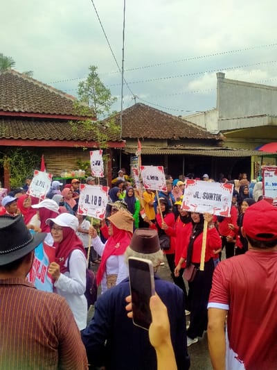 The image depicts an outdoor procession or gathering occurring on a paved street in Mojogedang, Indonesia, under an overcast sky. A large crowd, primarily composed of adults, fills the foreground and mid-ground. Many participants wear red tops or shirts, often combined with white garments. Several individuals, including women wearing hijabs, are holding aloft white rectangular signs mounted on poles. Prominent text visible on these signs includes "JL. SUNTIK" (Jalan Suntik), "JL. IUD" (Jalan IUD), and "JL. PIL" (Jalan Pil), accompanied by red graphic elements, indicating a focus on health or family planning initiatives.
In the immediate foreground, a man in a dark brown striped shirt and a black hat is partially visible on the left. Centrally, a man wearing a dark blue outfit and a brown traditional cap is holding a black smartphone vertically with his right hand, pointed towards the procession, indicating recording activity. Another hand in the lower center also holds a smartphone. To the right, the back of a person in a red shirt with white trim is visible.
The background features typical Indonesian architecture, including two traditional houses with brown tiled roofs on the left, one with red-painted lower walls. A sparse tree stands between these structures. Further right, a lighter-colored, multi-story building with a more modern design is present. Overhead, multiple utility lines and poles traverse the scene. An Indonesian flag (red and white) is partially visible in the mid-ground. In the far background, remnants of a vendor's stall with a red canopy and other items are discernible, suggesting a community or market setting.